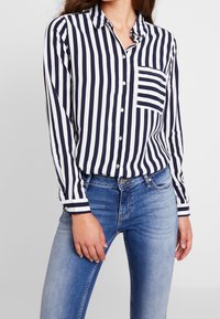 Chemise à rayures bleu marine et blanches avec col, une poche poitrine unique et manches longues. Associée à un jean en denim bleu.