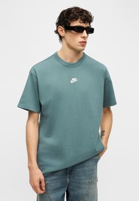 Νεαρός άνδρας που φοράει μαύρα ορθογώνια γυαλιά ηλίου, μπλε-πράσινο t-shirt Nike και μπλε τζιν, στέκεται με το ένα χέρι στην τσέπη.