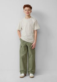 Lichtbeige t-shirt met korte mouwen, olijfgroene cargobroek met zijzakken, witte sneakers met beige accenten, staand tegen een effen achtergrond.