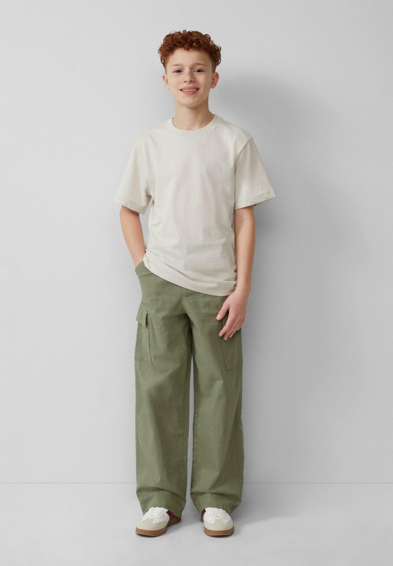 Lichtbeige t-shirt met korte mouwen, olijfgroene cargobroek met zijzakken, witte sneakers met beige accenten, staand tegen een effen achtergrond.