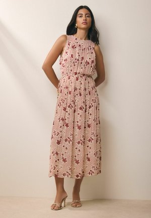 Žena stojaca, oblečená v béžových šatách bez rukávov po polovicu lýtka s červeným kvetinovým vzorom a zlatými sandálmi na vysokom opätku na jednofarebnom pozadí.