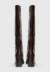 Bottes en cuir marron jusqu'au genou avec une texture lisse, des talons rectangulaires et des zips discrets sur les côtés intérieurs.