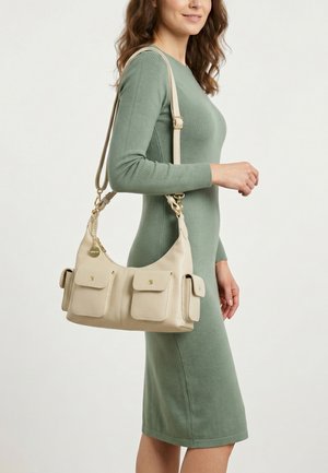 Vrouw in een groene jurk met lange mouwen draagt een beige handtas met meerdere voor- en zijzakken over haar schouder.