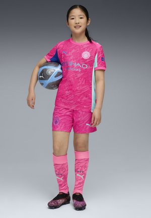 Conjunto de fútbol rosa que incluye una camiseta con un diseño, pantalones cortos y calcetas hasta la rodilla. La indumentaria incluye botas negras y multicolores, sosteniendo un balón azul.