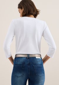 Vit kortärmad skjorta med trekvartslång ärm, parat med mörkblå denimjeans och ett texturerat silverbälte. Bakgrunden är neutral.