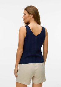 Top a coste di un blu navy con bordi ondulati, caratterizzato da uno scollo a V sul retro, abbinato a pantaloni corti beige chiaro. Texture a maglia liscia.