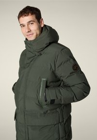Uomo che indossa un giubbotto invernale imbottito verde scuro con colletto alto, mani in tasca, su uno sfondo beige uniforme.