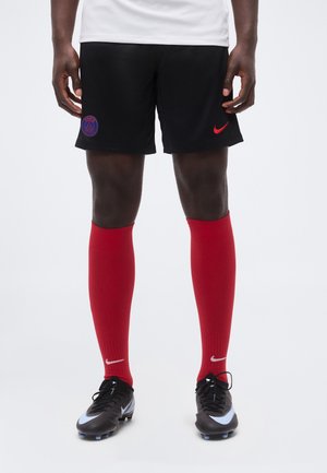 Jambes portant un short noir du Paris Saint-Germain, des chaussettes rouges Nike montantes et des chaussures de football Nike noires sur fond blanc.