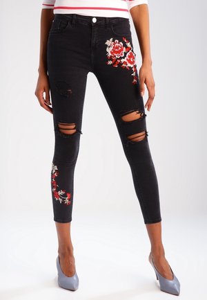 Woman wearing black ripped skinny jeans with red and white floral embroidery, gray high heels, and a white shirt with red stripes.