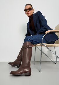 Bruine leren laarzen tot de knie met een slank ontwerp, voorzien van een driehoekig accent en een lage gestapelde hak. Gladde textuur met een gepolijste afwerking.