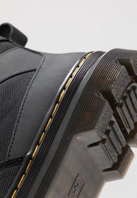 Gros plan sur la semelle noire d'une chaussure avec un motif de semelle profond, une couture jaune le long du bord, et une tige en tissu texturé.