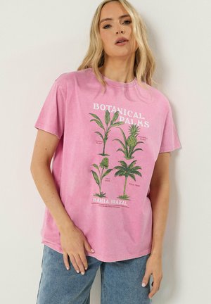 Kobieta ubrana w różowy T-shirt z zielonymi botanicznymi ilustracjami palm i tekstem, w połączeniu z jasnoniebieskimi dżinsami, stojąca na tle białej ściany.