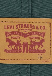 Tan label značky Levi Strauss & Co. s červeným motivem koní a kalhot, "Kvalitní oblečení," velikost 12M, přišitá na okraji.