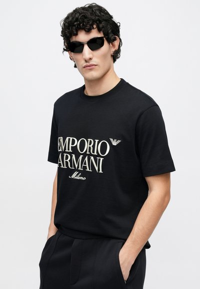 Μαύρο βαμβακερό T-shirt με το λογότυπο «EMPORIO ARMANI» σε ανοιχτό μπεζ, με κοντά μανίκια και στρογγυλή λαιμόκοψη. Το μοντέλο φοράει μαύρα γυαλιά ηλίου και σκούρο παντελόνι.
