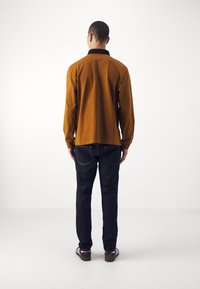 Hnědá košile s dlouhým rukávem a černým límečkem, kombinovaná s tmavými džínami. Tkanina působí hladce, s volným střihem a kontrastním prošíváním.