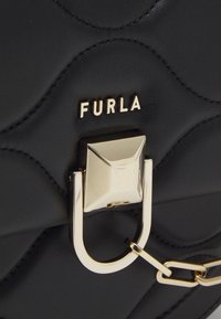 Borsa a mano in pelle nera con texture trapuntata, accessori in oro, con una fibbia quadrata prominente e il nome del marchio "Furla" in rilievo.