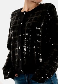 Cardigan noir avec une finition texturée, orné de sequins argentés en motif à carreaux et doté de grands boutons ronds.
