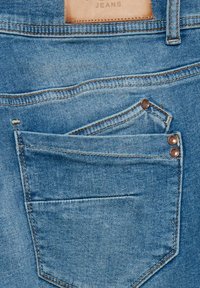 Los vaqueros de mezclilla azul cuentan con un bolsillo trasero con detalles de costura y un bolsillo para monedas más pequeño y en ángulo. Dos remaches de metal marrón acentúan el diseño.