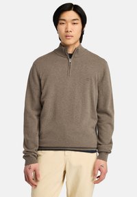Maglione zip-up marrone con colletto alto, polsini e orlo a costine. Texture morbida e branding minimale sul petto. Indossato sopra una maglietta scura.