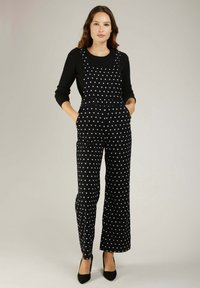 Combinaison salopette noire à pois avec des jambes larges, dotée de bretelles ajustables et de poches latérales. Portée avec un haut à manches longues côtelé noir.