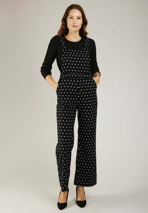 Combinaison salopette noire à pois avec des jambes larges, dotée de bretelles ajustables et de poches latérales. Portée avec un haut à manches longues côtelé noir.
