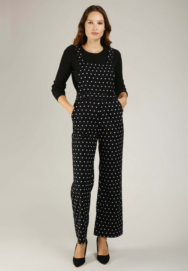 Combinaison salopette noire à pois avec des jambes larges, dotée de bretelles ajustables et de poches latérales. Portée avec un haut à manches longues côtelé noir.