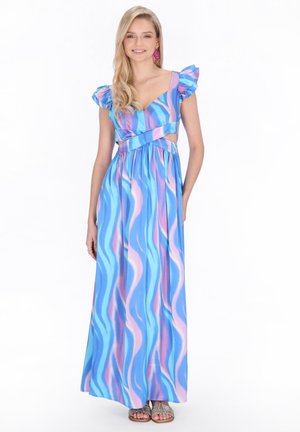 Donna sorridente, che indossa un abito lungo con motivo blu e rosa, ondeggianti, maniche a sbuffo e inserti laterali, in piedi con una mano dietro la schiena.