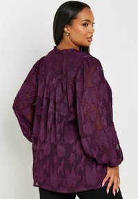 Blusa violacea trasparente con ricamo floreale testurizzato, maniche lunghe e dettaglio plissettato sulla schiena. Presenta un collo alto e una vestibilità ampia.