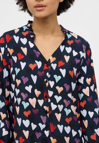 Button-up shirt featuring a navy background with a multi-colored heart pattern, ruffled collar, and black buttons. Soft fabric texture.
