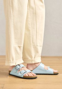 Svetlo modri sandali na odprti nos, z dvojno zaponko, plute podplat in črna gumijasta podloga, v kombinaciji s kremnimi hlačami širokega kroja.