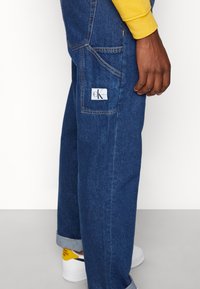 Blå denimjeans med en avslappnad passform, med en bakfickan som har en CK Jeans-etikett. Bena är uppvikta och de är kombinerade med vita sneakers.