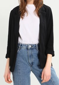 Černé sako z texturované látky s klopami s výstřihem a ohrnutými rukávy, doplněné bílým tričkem a modrými vysokými džínami.