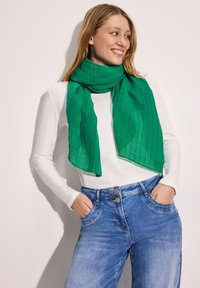 Grön plisserad scarf av lättviktsmaterial som hänger elegant runt nacken. Bärs tillsammans med en vit långärmad skjorta och blå jeans.