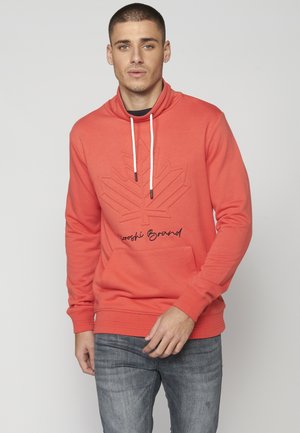 Koralikowy hoodie z kieszenią kangurką, sznurkiem przy szyi, wytłaczaną grafiką liścia i haftowanym logo, zestawiony z szarymi dżinsami.