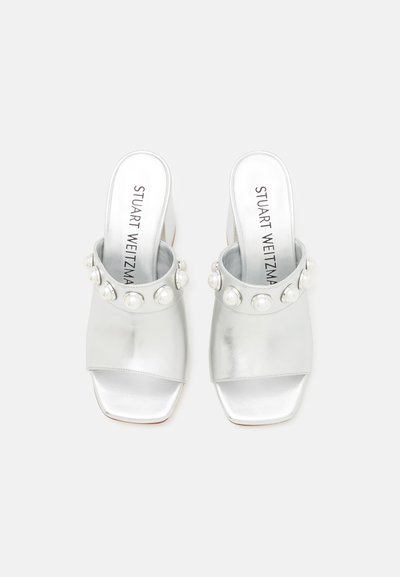 Mules métalliques argentés à talons hauts avec un bout carré, une large lanière et des accents de perles décoratives autour de la cheville. Texture lisse.