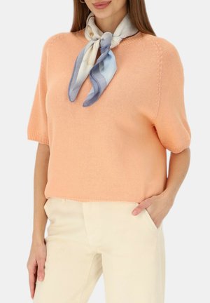 Kobieta w brzoskwiniowej, krótkorękawowej dzianinowej bluzce, kremowych wysokich spodniach i niebiesko-białej jedwabnej apaszce zawiązanej wokół szyi.