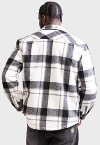 Camicia in flanella a quadri bianca e nera, con colletto, polsini con bottoni e un tessuto morbido e strutturato. Il design include grandi quadrati a contrasto.