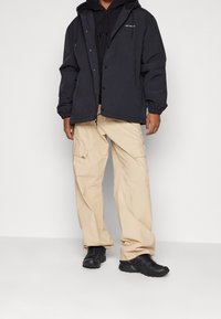 Man som bär svart huva, svart hoodie, beige cargo-byxor och svarta sneakers, står mot en enkel vit bakgrund.