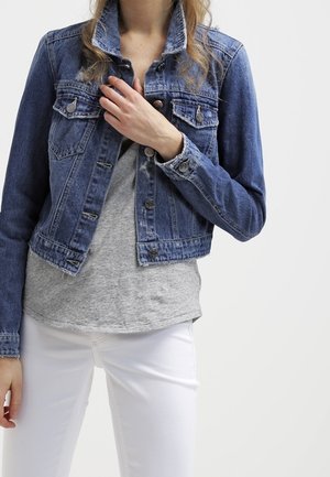 Person iført en blå denimjakke over en grå skjorte og hvide bukser, med en hånd der justerer jakke kraven.