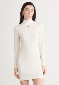 Rochie tricotată cu mânecă lungă, de culoare crem, cu guler înalt și un logo discret al brandului la piept. Textură netedă și siluetă ajustată.
