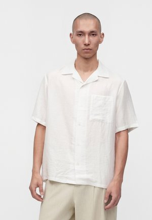Homme à la tête rasée portant une chemise blanche ample à manches courtes et un pantalon beige sur un fond clair uni.