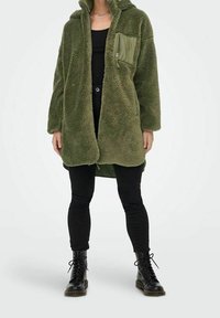 Zelený fleecový kabát s kapucí, kapsou na prsou na zip a přehnaným střihem, doplněný černými legínami a bojovými botami. Jemná textura.