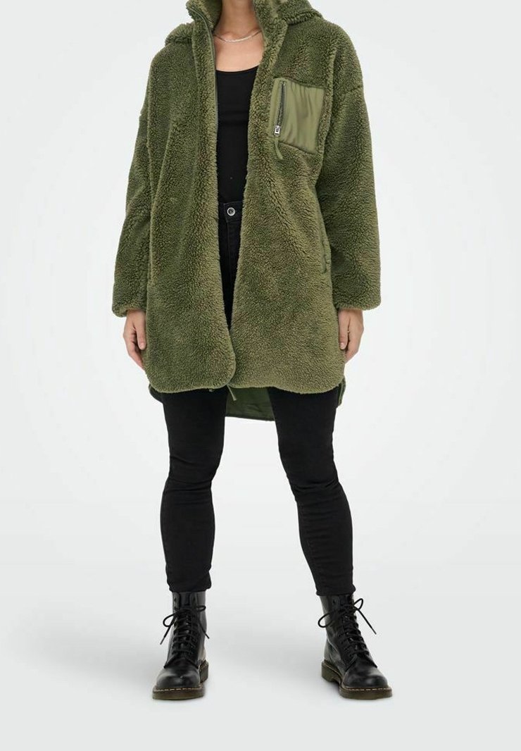 Zelený fleecový kabát s kapucí, kapsou na prsou na zip a přehnaným střihem, doplněný černými legínami a bojovými botami. Jemná textura.