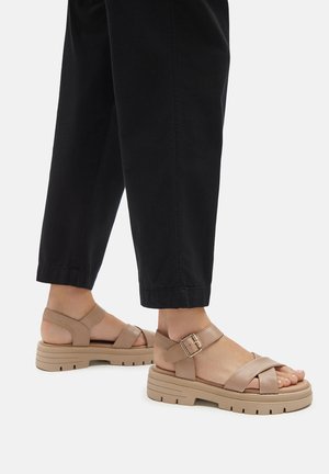 Bež kožne sandale s dizajnom križnog remena, podešivom kopčom i debelim, brušenim đonom, uparenim s crnim skraćenim hlačama.