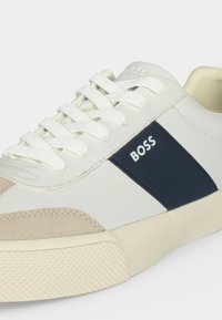 Baskets blanches à lacets avec détail en daim beige et panneau bleu marine portant le mot BOSS sur le côté, et semelle crème texturée.