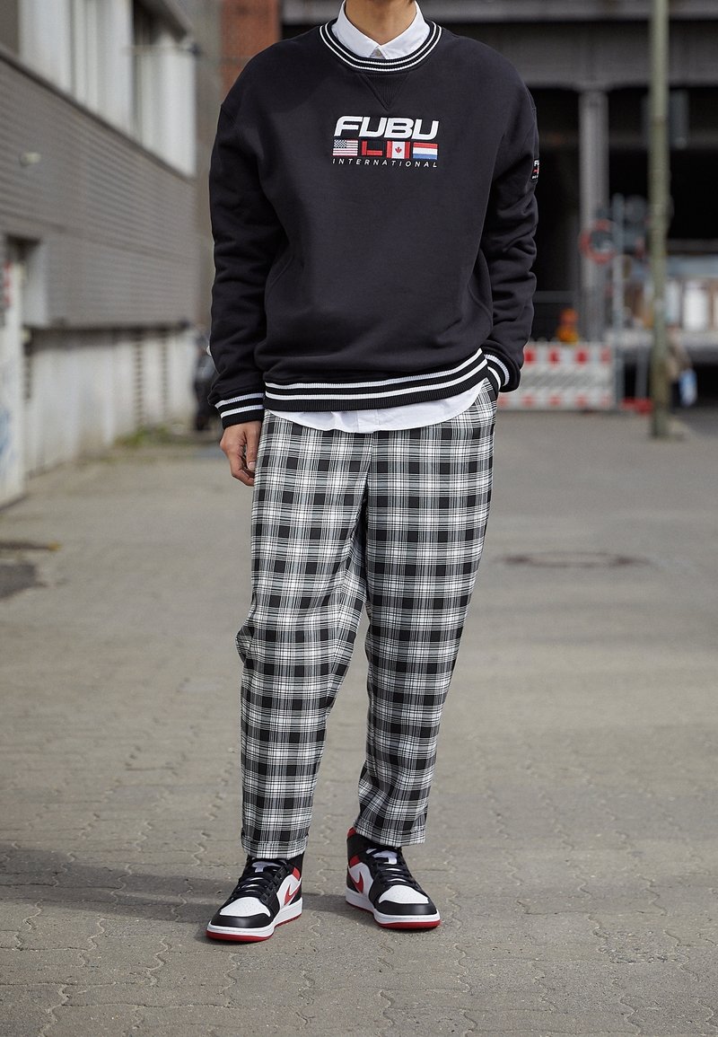 Černá mikina s bílými pruhovanými manžetami, se červeným a modrým logem. Šedé kostkované kalhoty a černé/červené tenisky dotvářejí outfit.