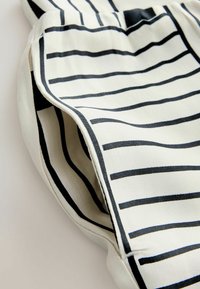 Približeni prikaz kremaste tkanine s crnim horizontalnim i vertikalnim prugama, prikazujući šavove i slojevite nabore.