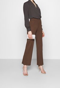 Pantalon marron à jambes larges associé à un chemisier foncé et des talons à brides. La tenue est complétée par une petite pochette argentée ornée de décorations.
