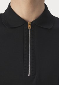 Gros plan d'une chemise noire à col avec une fermeture éclair verticale argentée et un tire-zip doré au centre de l'encolure.