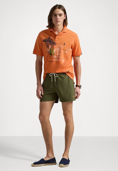 Camisa polo laranja com estampado gráfico, combinada com calções verdes-oliva. O modelo usa espadrilhes azul-marinho. Conjunto de verão simples e casual.